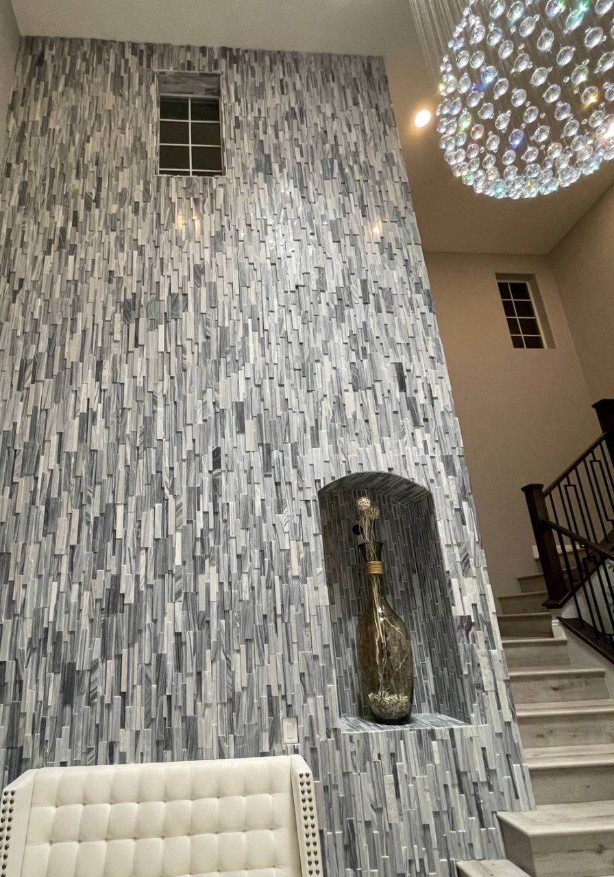 A two-story entry accent wall covered in grey and white split-face marble ledger strips, with a tiled arched niche holding a tall glass vase, a small window near the ceiling and a crystal chandelier